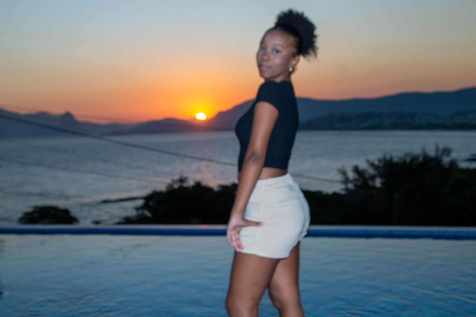 Mulher posa ao lado de uma piscina ao pôr do sol; montanhas e água ao fundo.