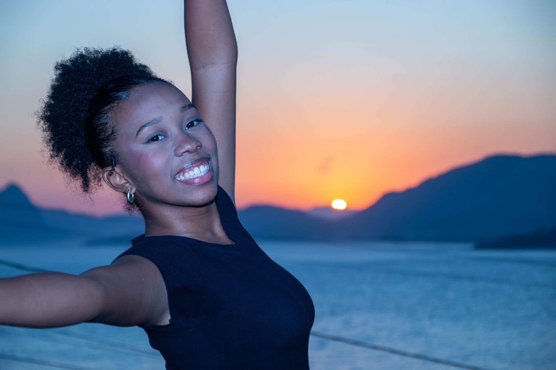 Mulher sorri, braços levantados, em frente ao pôr do sol sobre a água e as montanhas.