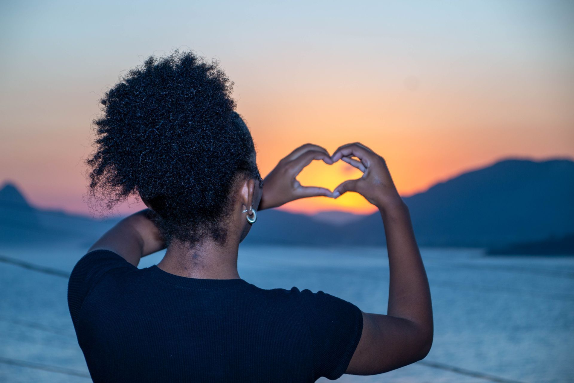 Pessoa fazendo um formato de coração com as mãos, emoldurando um pôr do sol sobre a água.