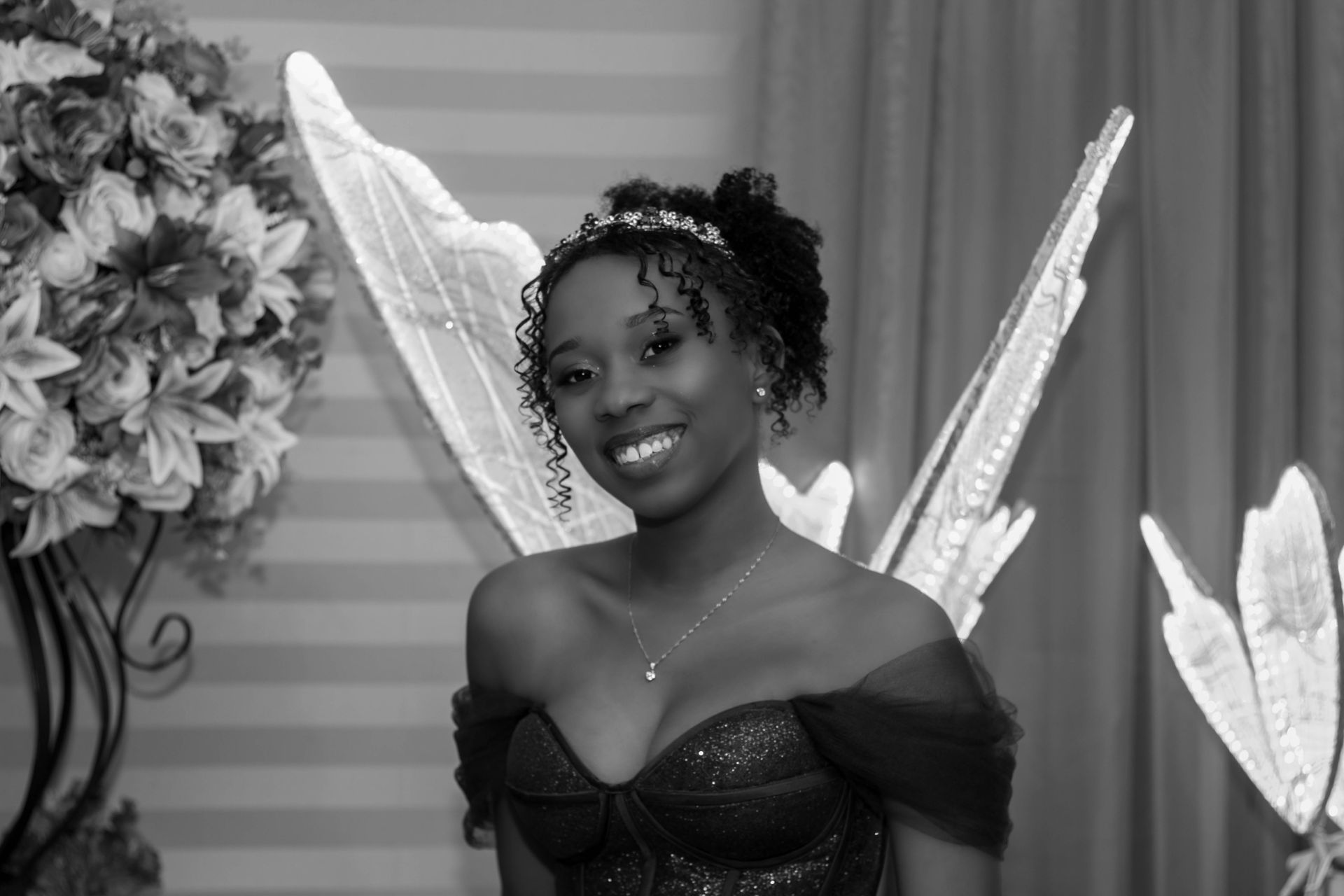 Mulher de cabelos escuros e cacheados, usando uma fantasia de fada, sorrindo. Ela tem um adorno de cabeça com joias e asas.