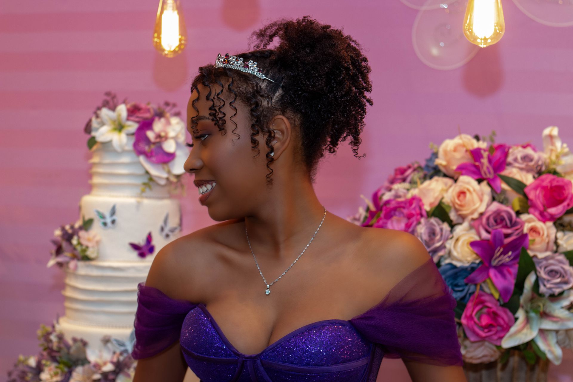 Jovem sorrindo, usando vestido roxo, em uma festa, perto de um bolo e flores.