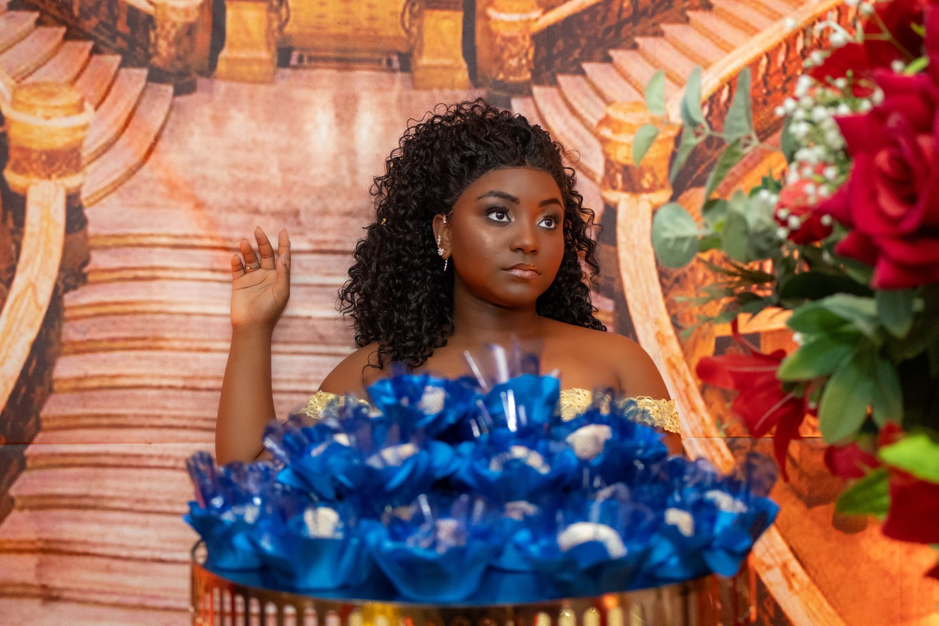 Mulher com cabelo cacheado posa ao lado de balas azuis, arranjos florais e uma escada ao fundo.