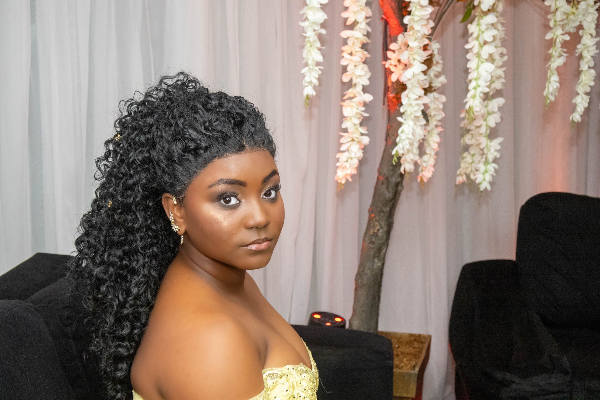 Mulher de cabelos pretos cacheados, usando um vestido amarelo, olha para a câmera, perto de decorações florais brancas.