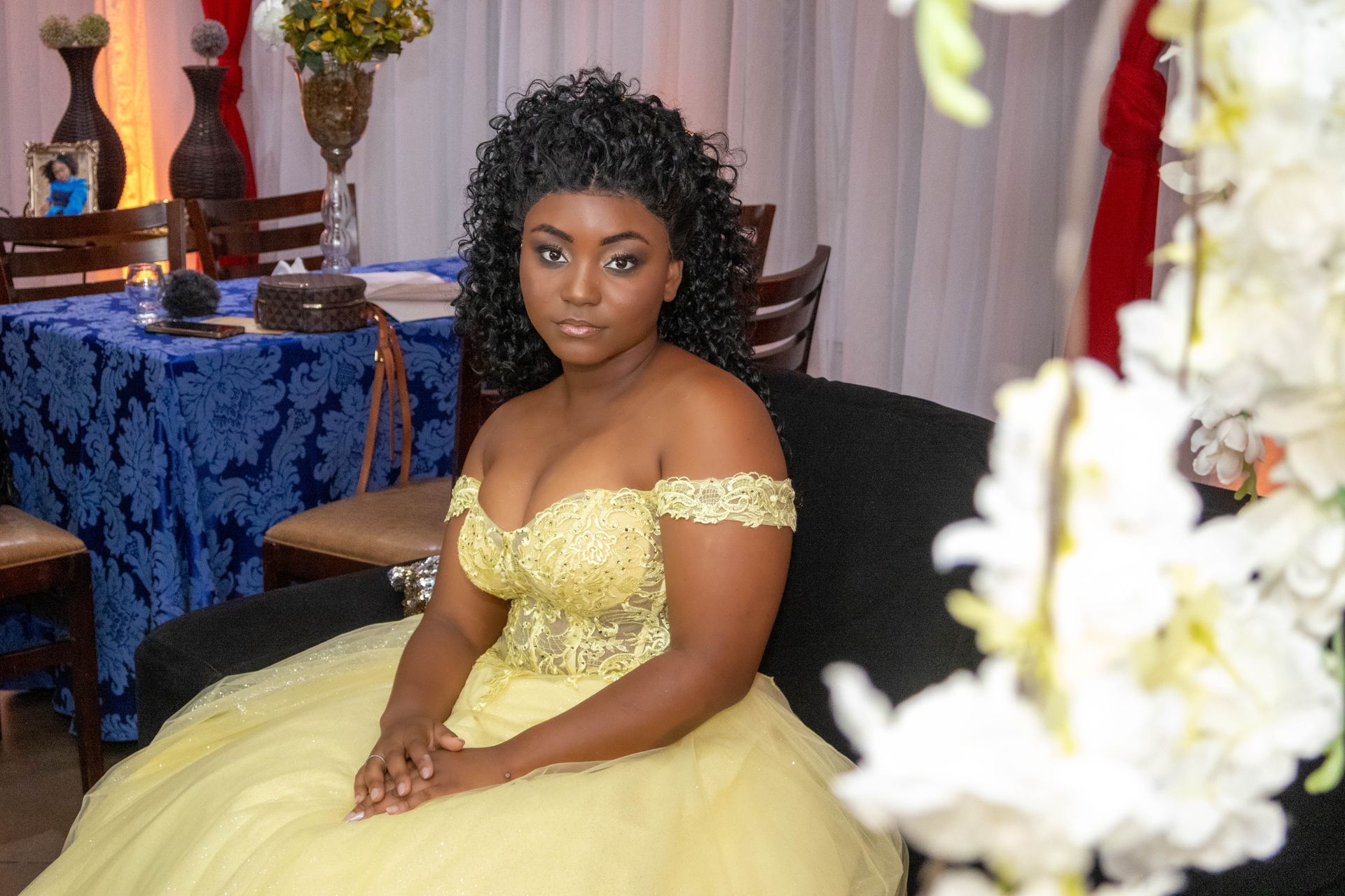 Mulher com um vestido amarelo tomara que caia, sentada em um sofá preto. Cenário formal para evento com flores brancas.