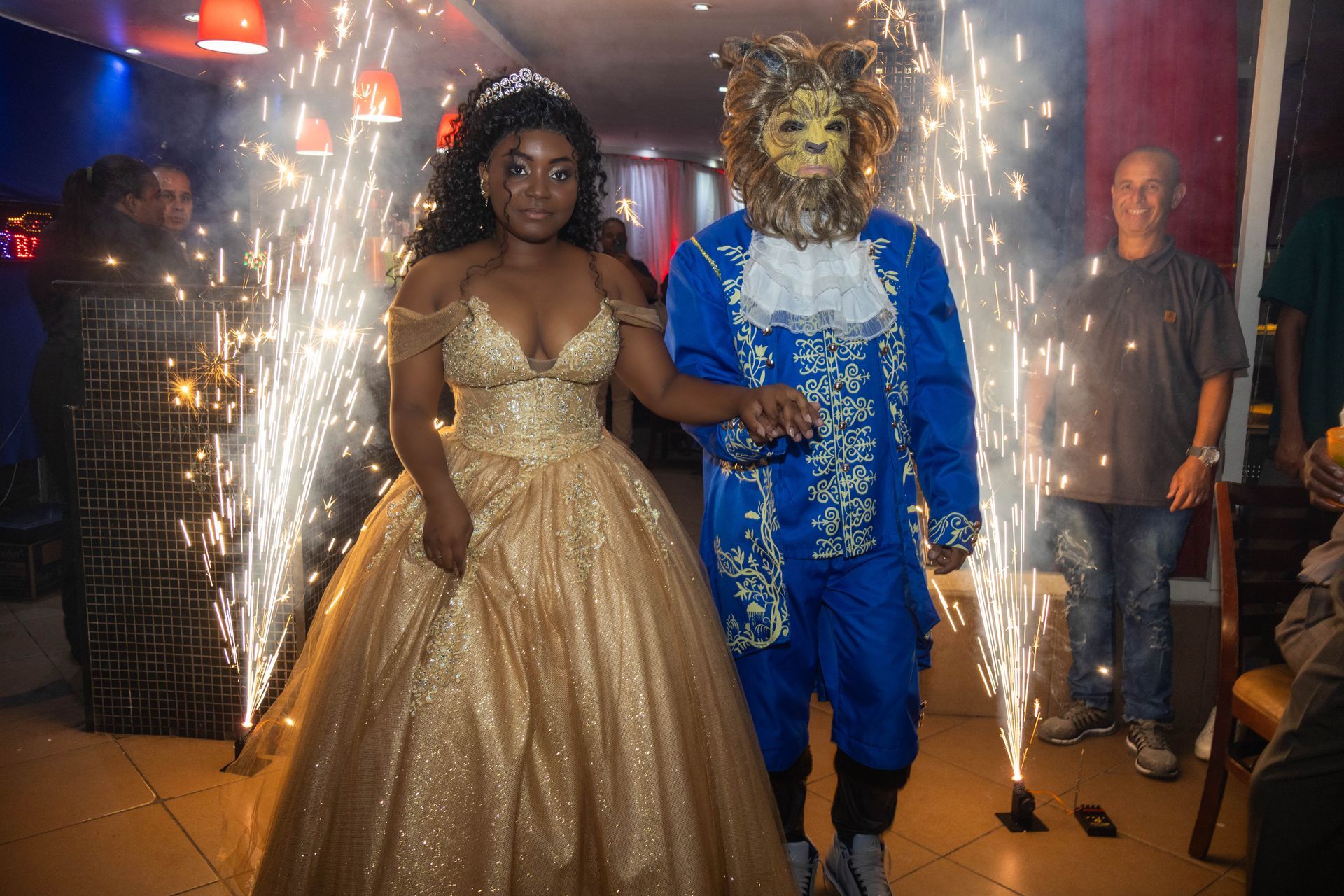 Mulher em vestido dourado caminha com pessoa com máscara de leão em meio a faíscas.