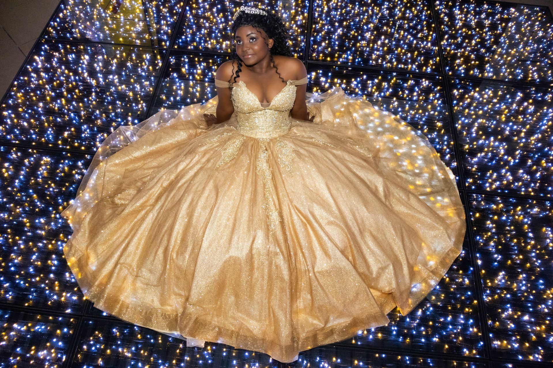 Mulher de vestido dourado na pista de dança iluminada, sorrindo e olhando para cima.