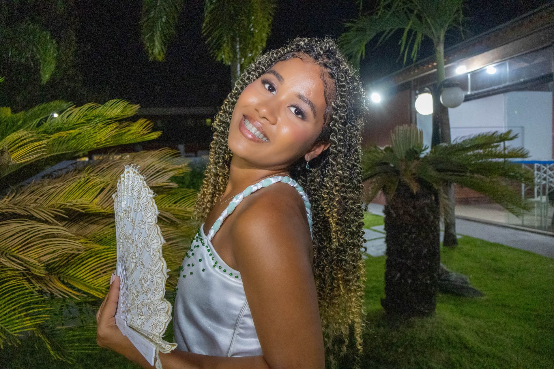 Mulher com cabelo trançado sorrindo, segurando uma bolsa brilhante; ao ar livre perto de vegetação.