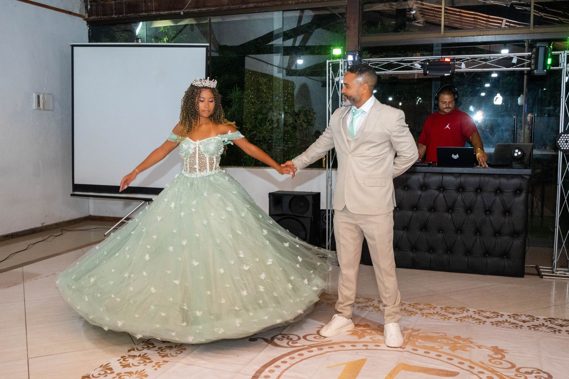 Mulher em vestido de baile verde menta dança com um homem de terno claro; ambiente de festa com DJ.