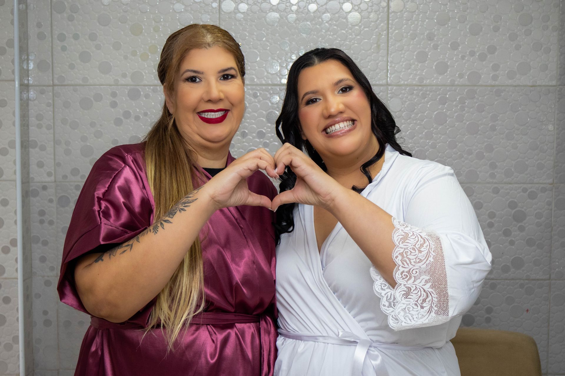 Duas mulheres de roupão fazendo formato de coração com as mãos, sorrindo.