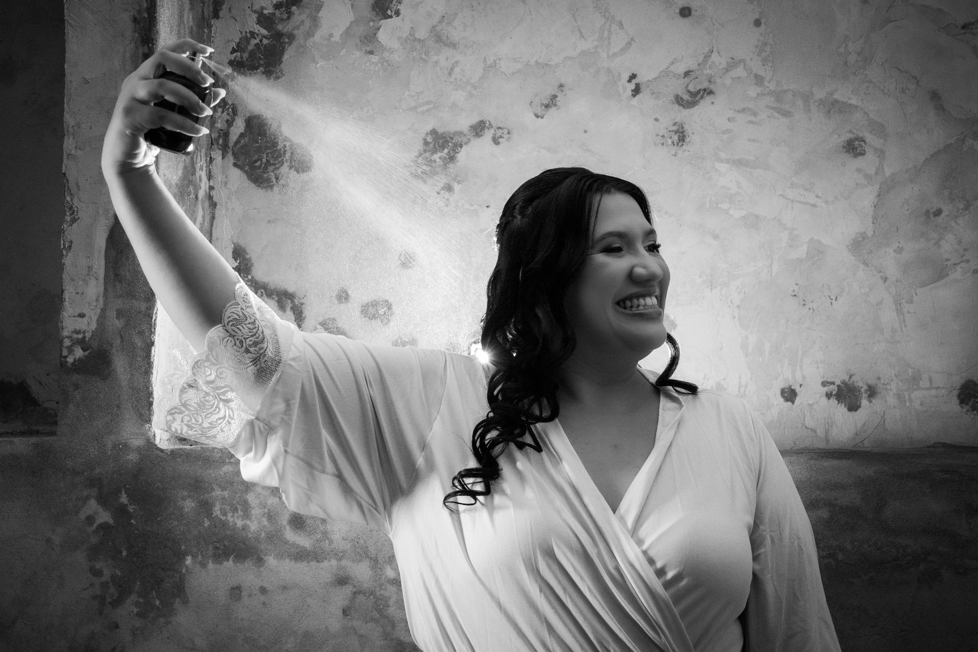 Mulher borrifa perfume, com o braço levantado, sorrindo, contra uma parede texturizada.