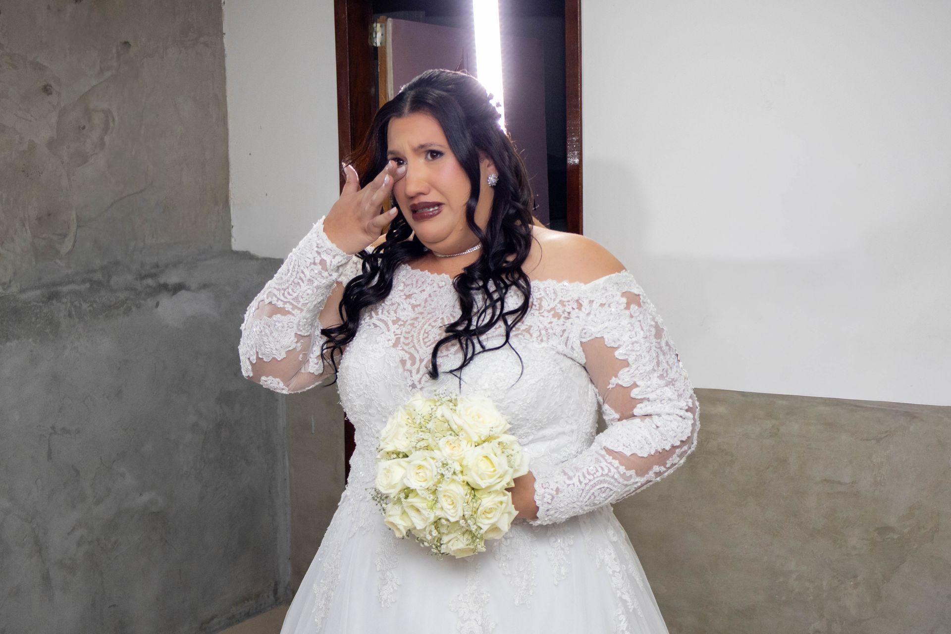 Noiva enxugando lágrimas, usando um vestido de noiva branco de renda e ombros à mostra, segurando rosas brancas, em ambiente interno.