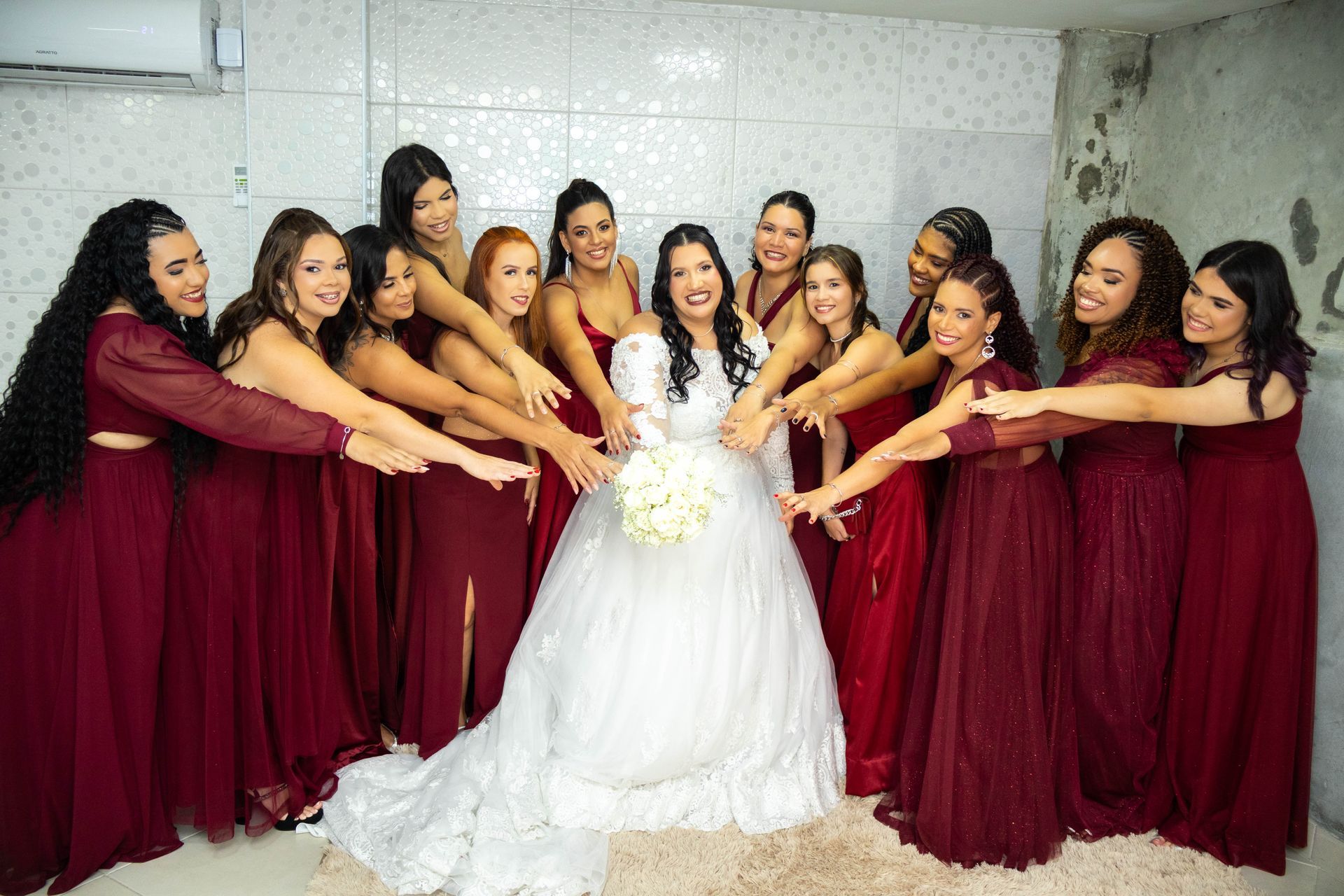 Noiva cercada por madrinhas, todas de mãos dadas em sua direção. Elas usam vestidos cor de vinho em um salão com paredes brancas.