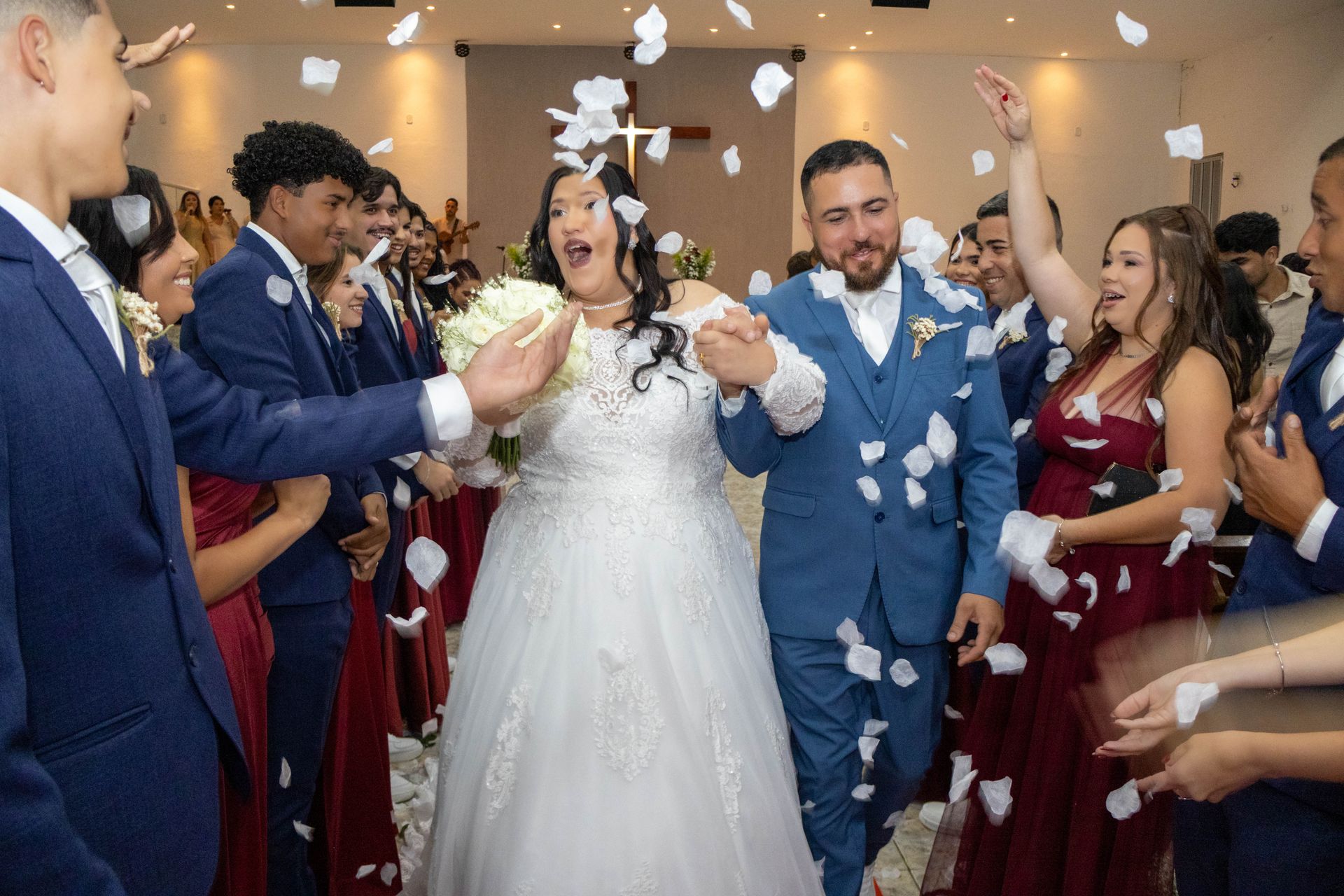 Recém-casados caminham por um arco enquanto convidados jogam pétalas, dentro de um local de casamento.