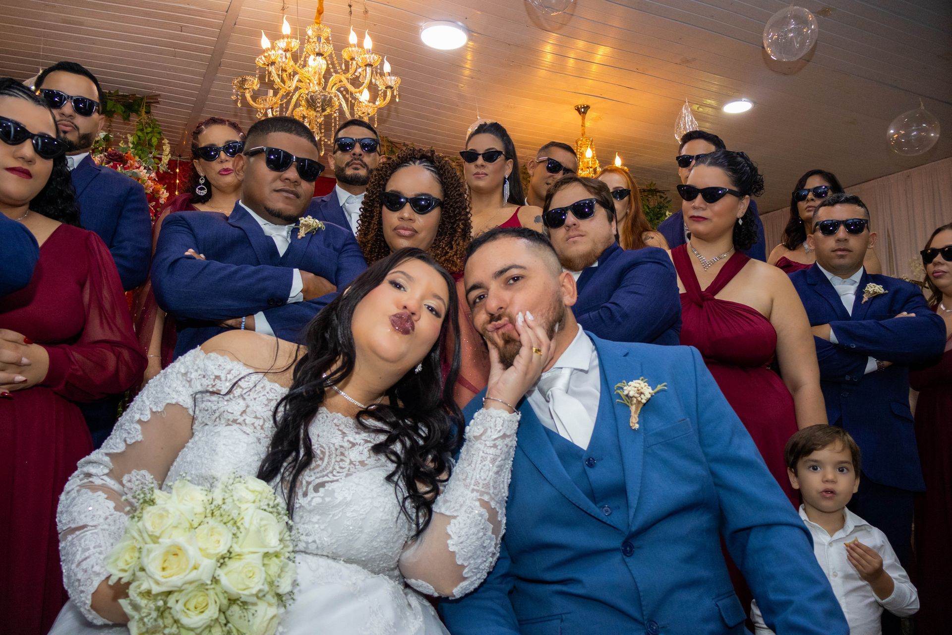 Recém-casados posam com seus convidados, todos usando óculos escuros e fazendo carinhas de beijo.