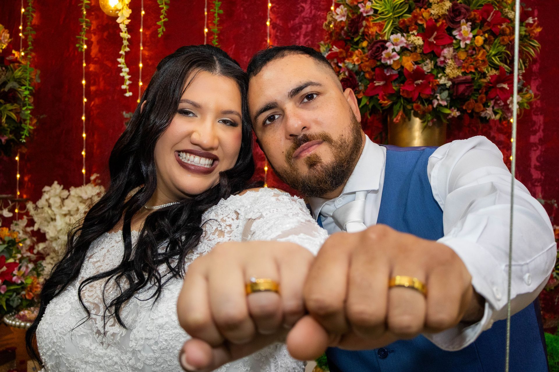 Noivos exibindo alianças, sorrindo para a câmera, com um fundo floral vermelho.