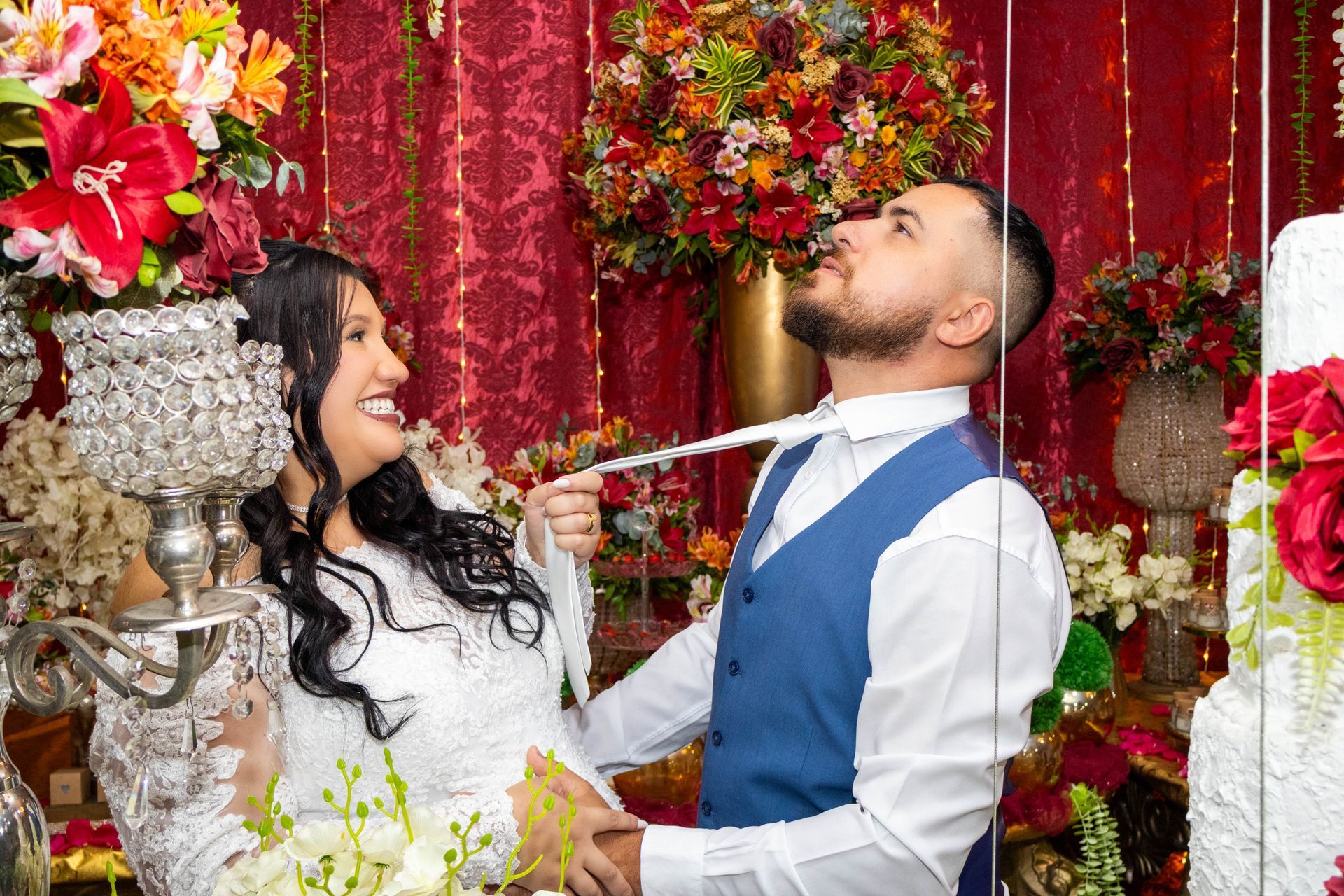 Noiva puxa a gravata do noivo de brincadeira. Eles sorriem, cercados por flores e bolo de casamento. Fundo vermelho.