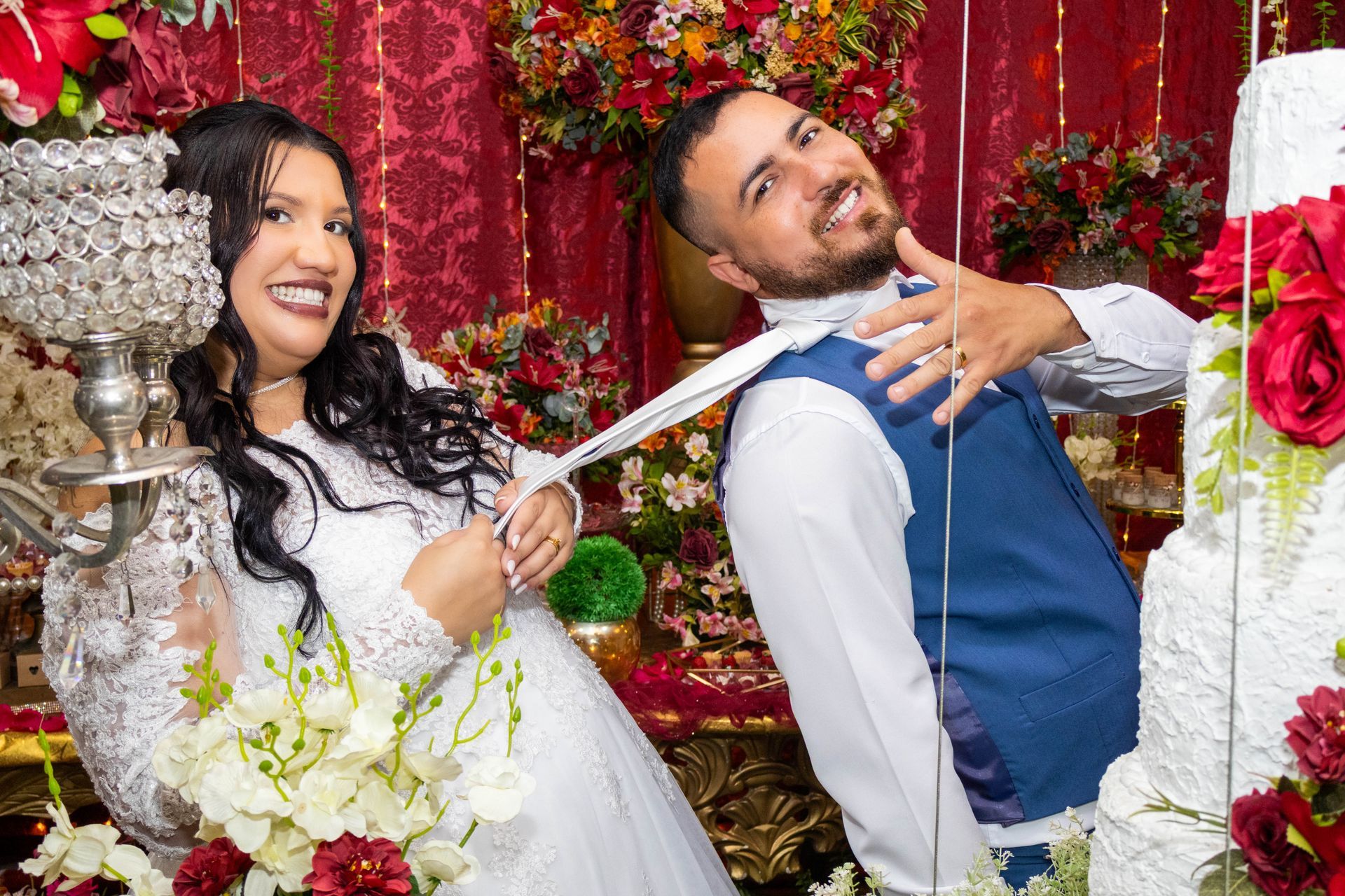Noiva brinca puxando a gravata do noivo, ambos sorrindo em uma recepção de casamento. Decoração floral vermelha e branca ao fundo.