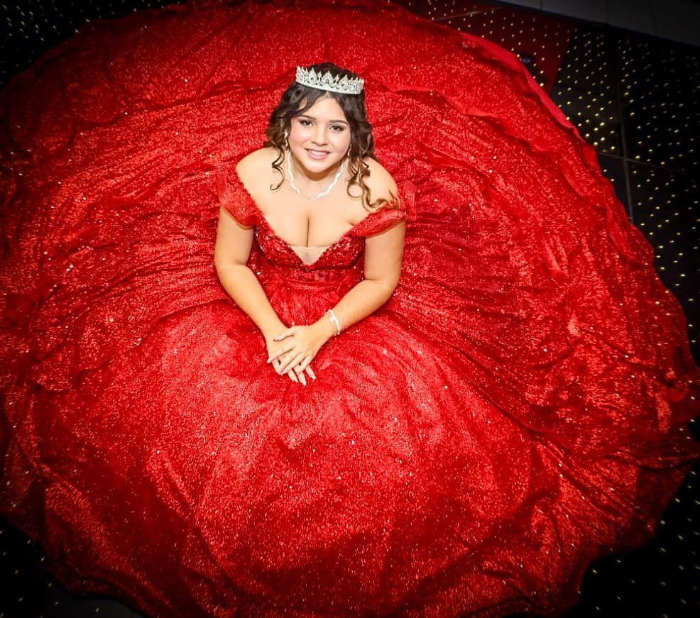 Mulher com um vestido de baile vermelho brilhante, tiara e colar, sentada em um chão estampado em preto e branco, sorrindo.