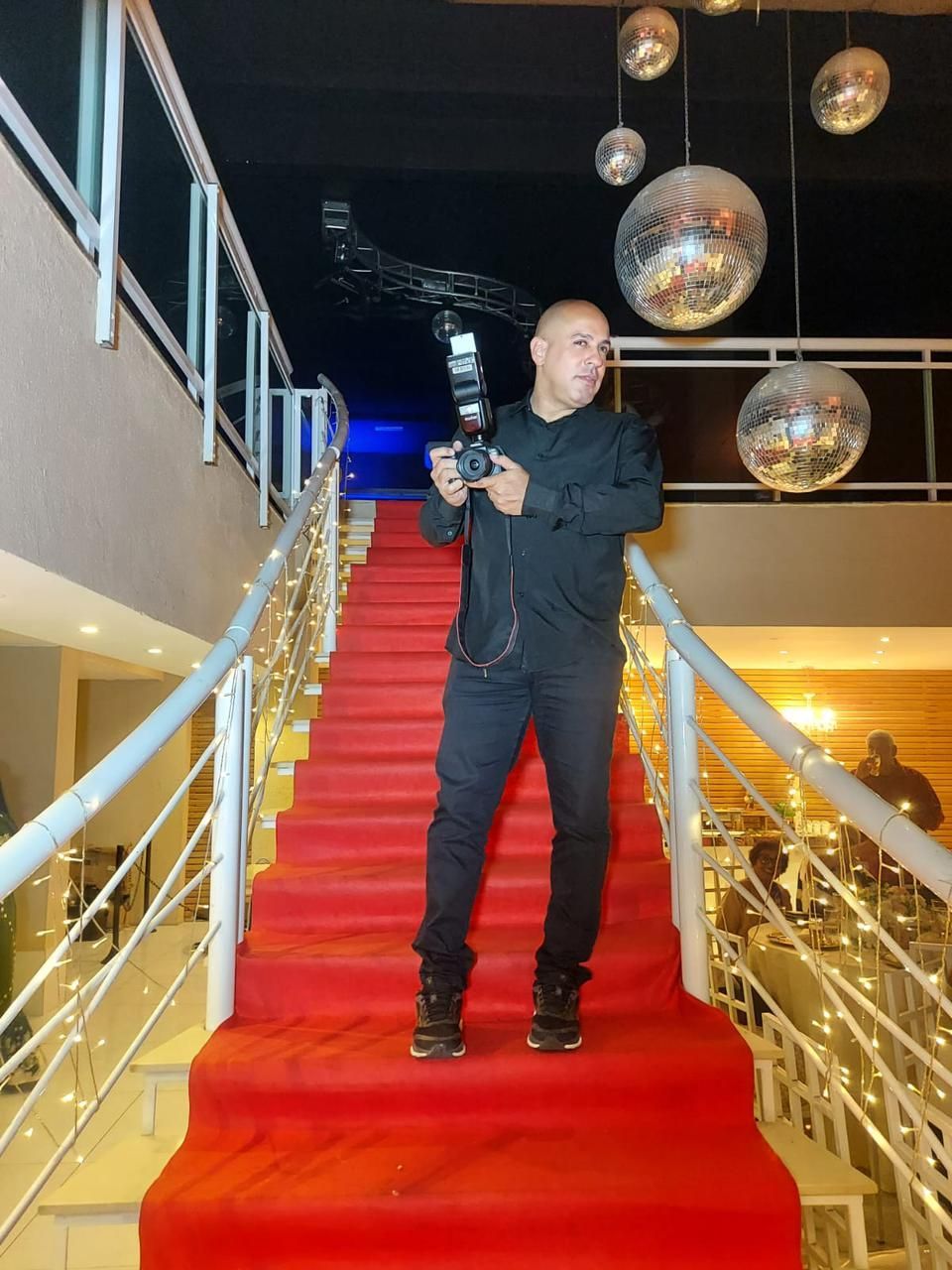 Homem com câmera em escada com carpete vermelho, segurando a câmera, olhando para a frente. Interior, luzes.