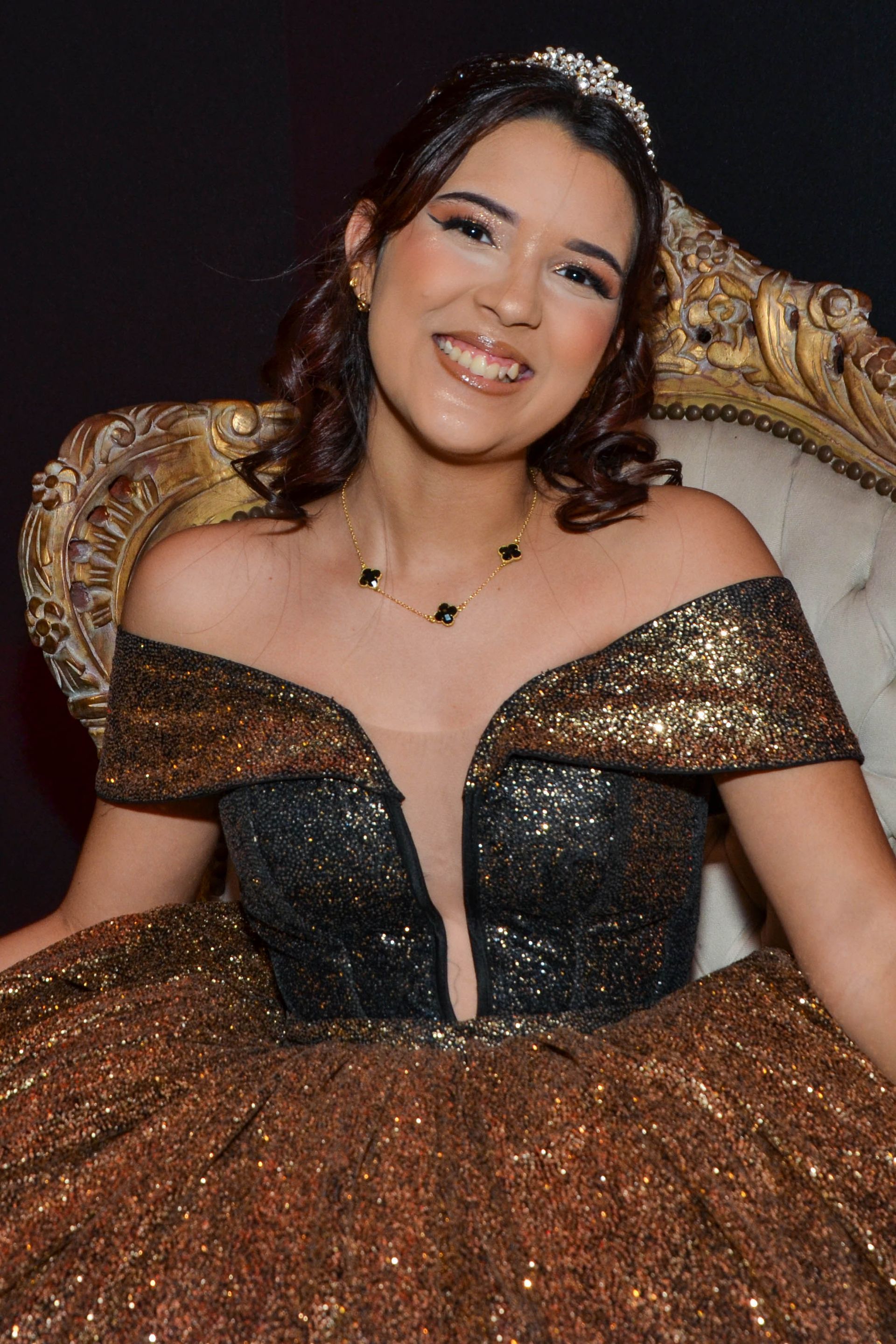 Mulher em vestido de lantejoulas douradas e pretas, sorrindo. Sentada em uma cadeira dourada ornamentada, usando tiara e colar.