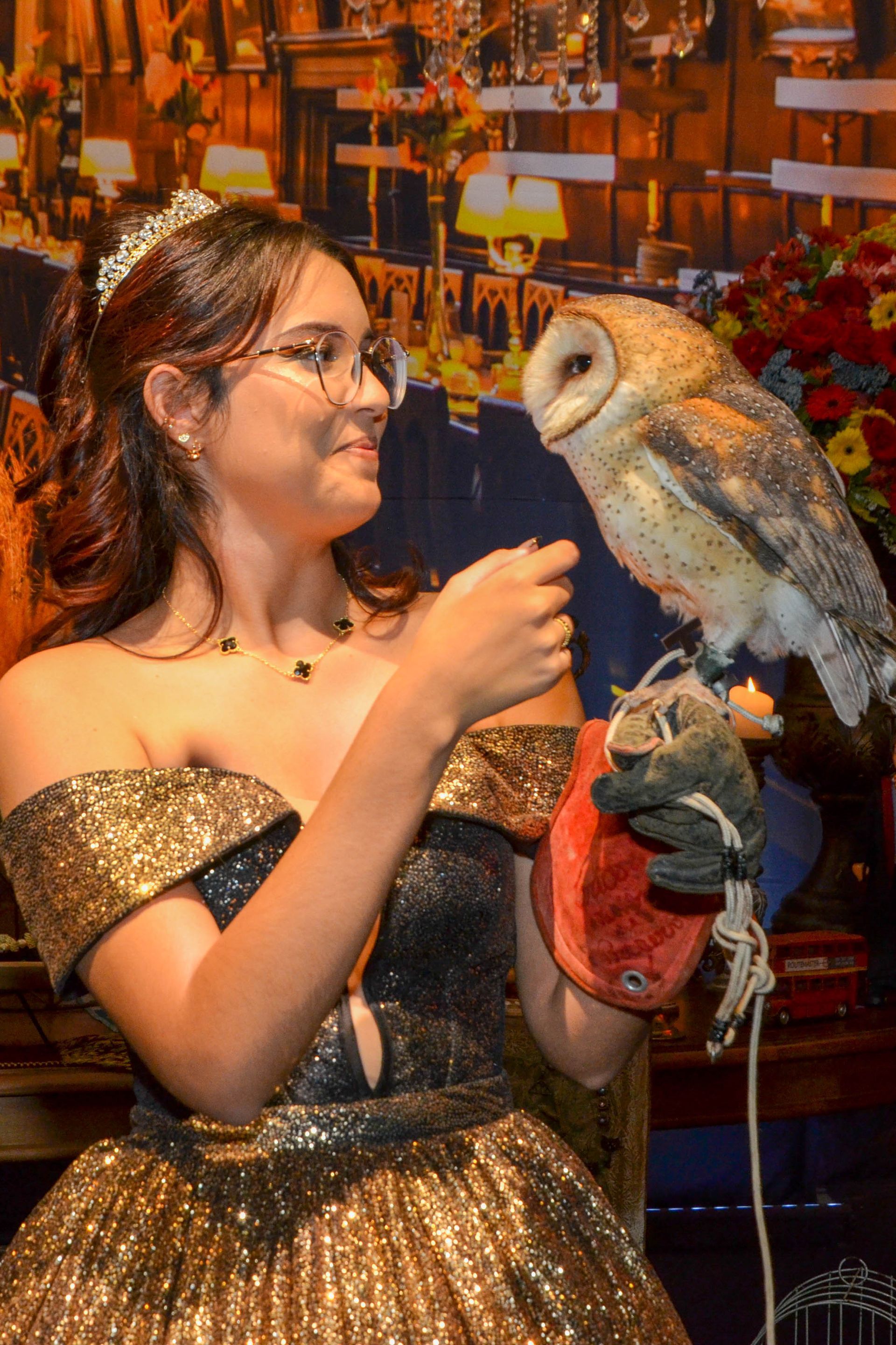 Mulher em vestido formal dourado e óculos segura uma coruja dentro de casa.