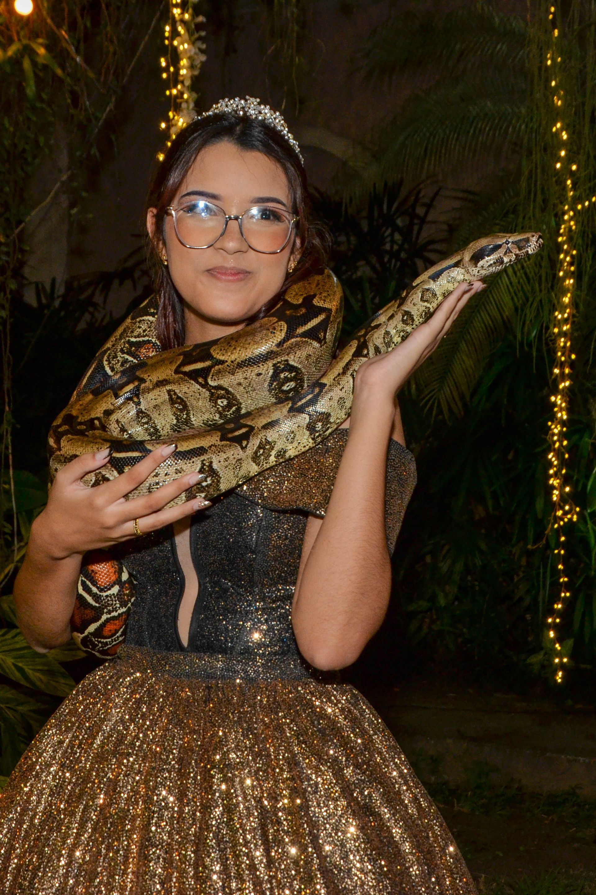 Mulher com um vestido brilhante segurando uma grande cobra no pescoço, sorrindo. Ao ar livre, iluminada com luzes de fada.