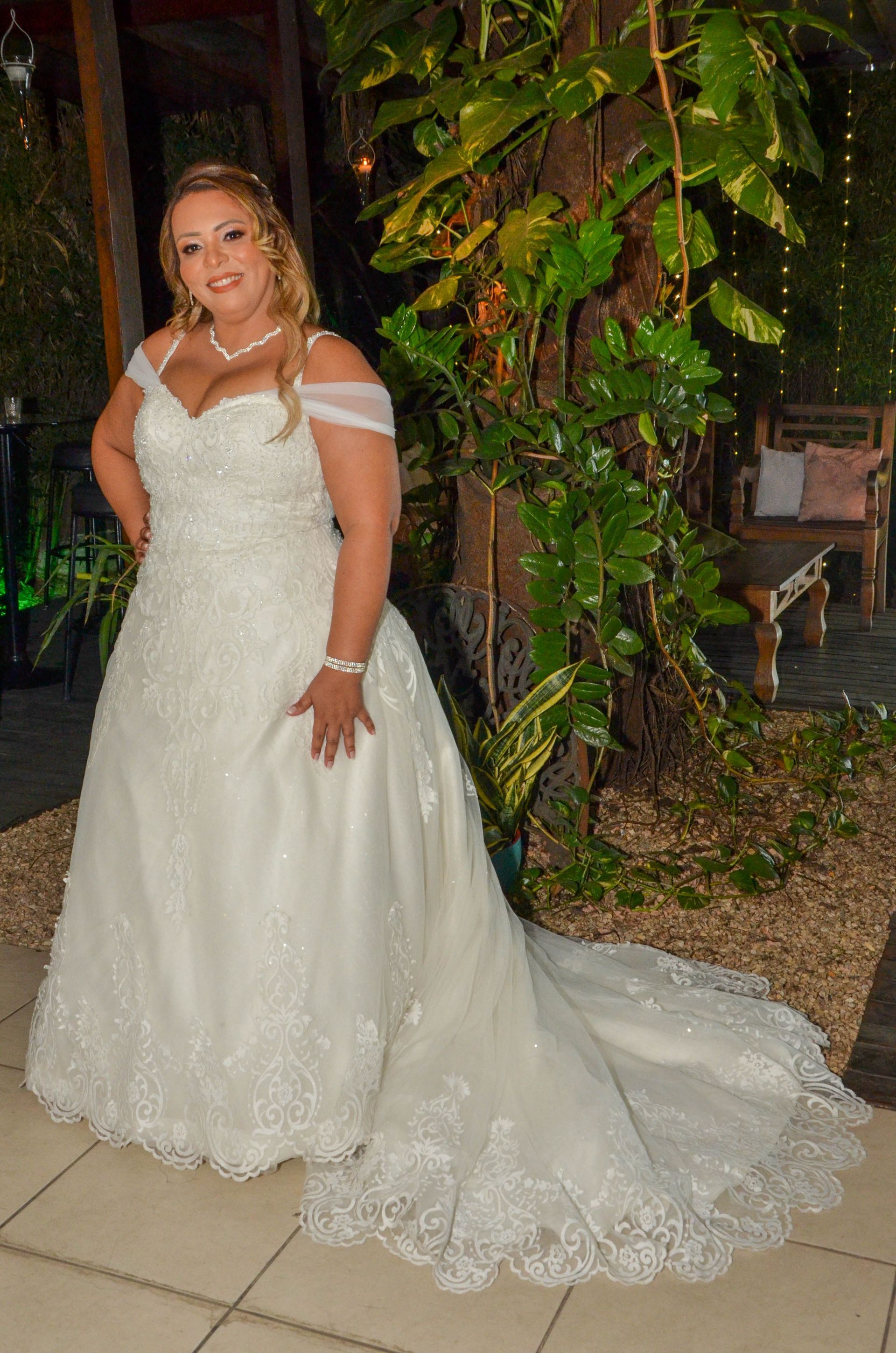 Mulher em um vestido de noiva branco sorri, parada perto de vegetação, um caminho de pedras e um banco de madeira.