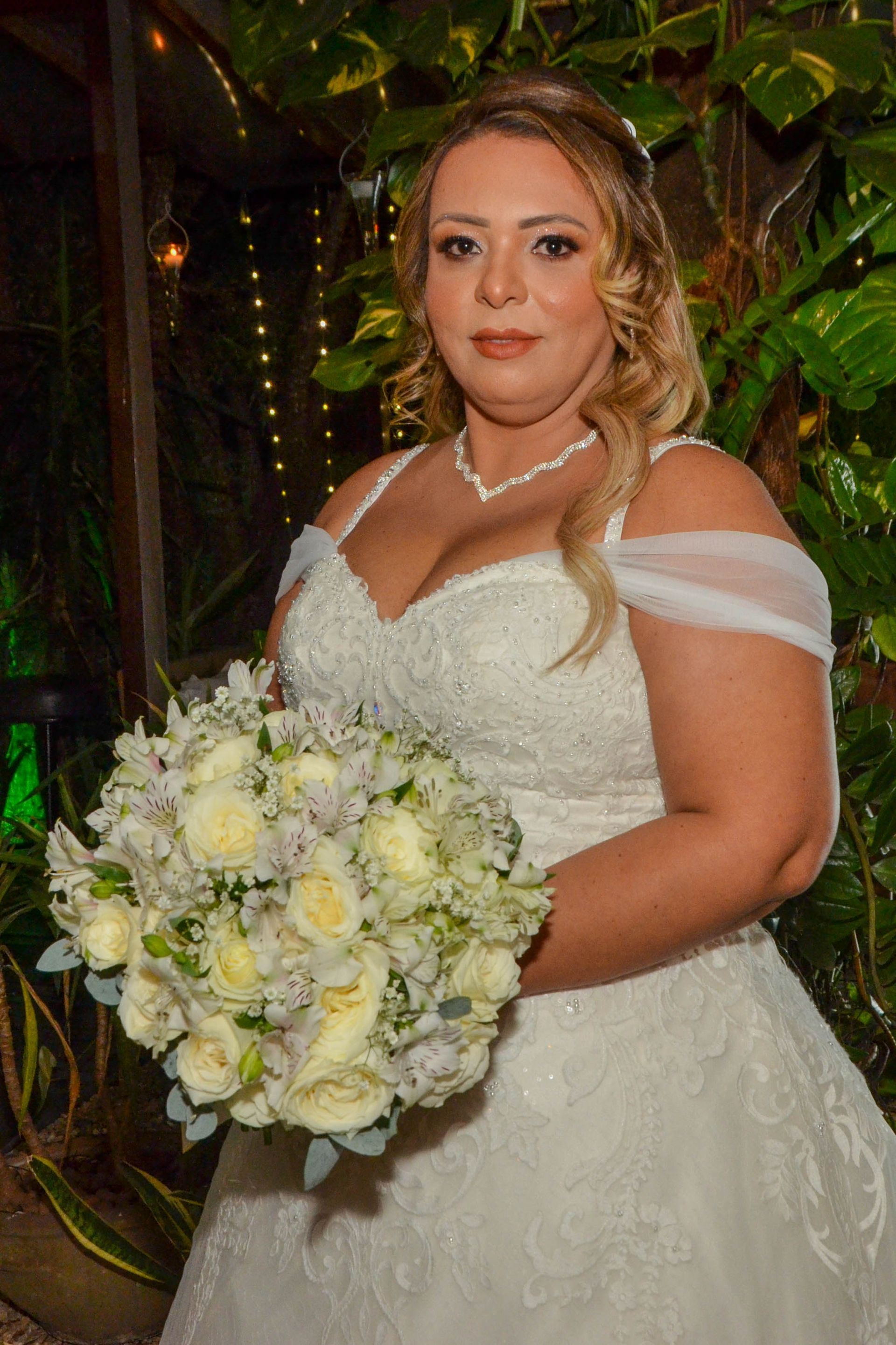 Noiva em um vestido branco com um buquê, em pé ao ar livre perto de uma área verde.