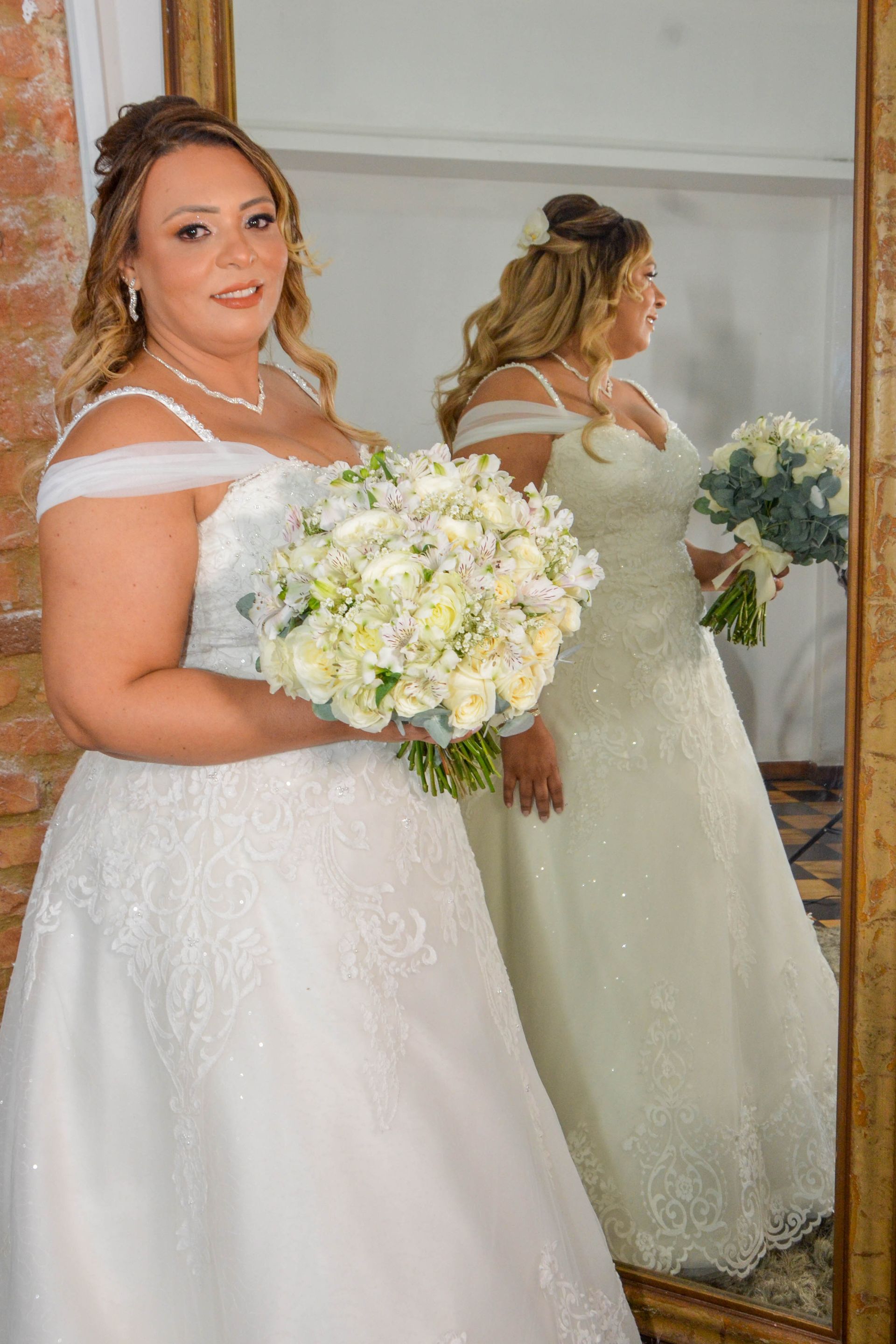 Noiva com vestido de noiva branco tomara que caia, segurando um buquê, em frente ao espelho.