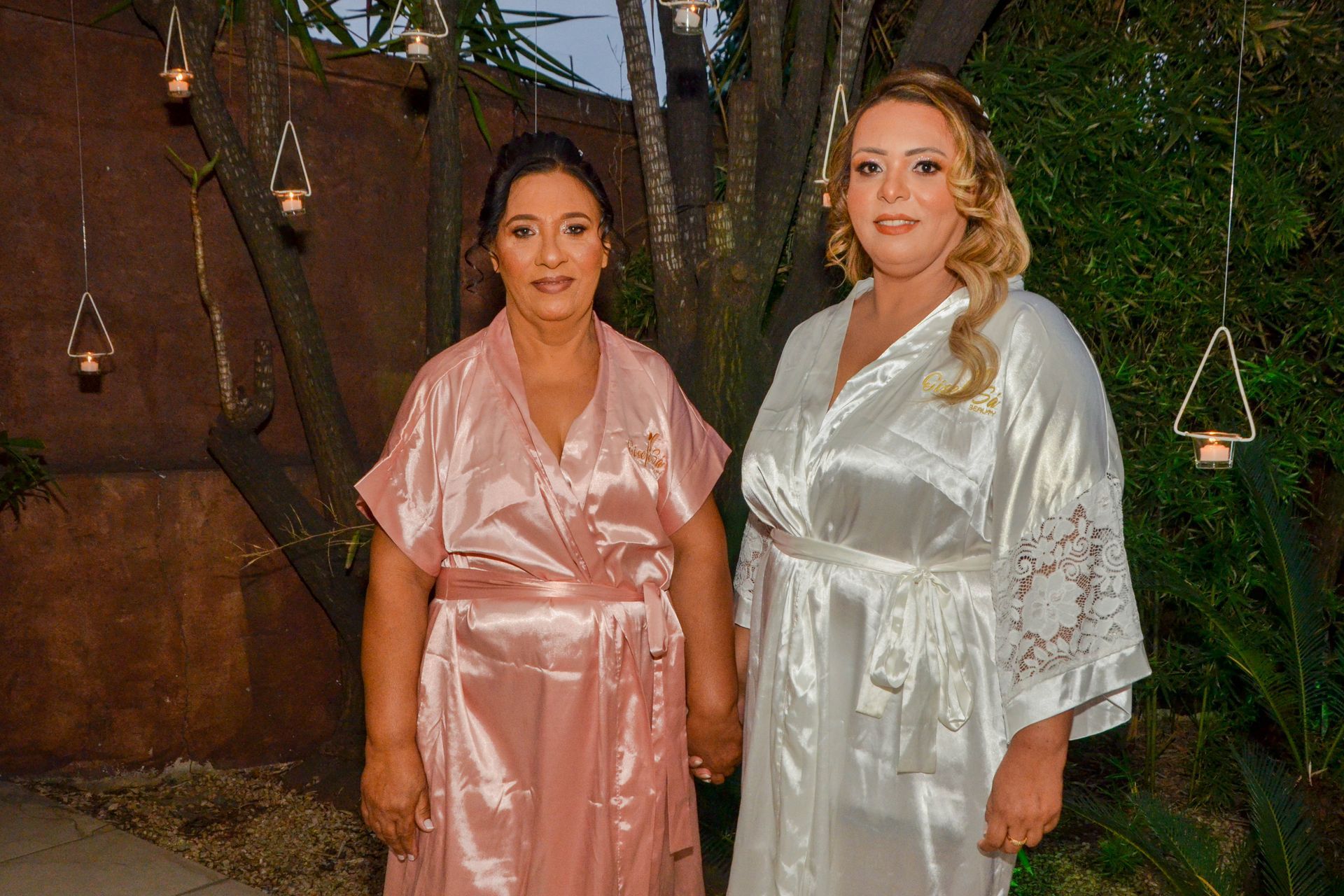 Duas mulheres de túnica estão do lado de fora, de mãos dadas. Uma veste rosa, a outra, branco. Castiçais pendurados ao fundo.