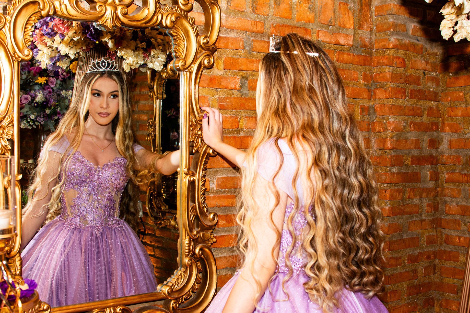 Mulher com vestido lilás e tiara olha para um espelho dourado ornamentado em frente a uma parede de tijolos.