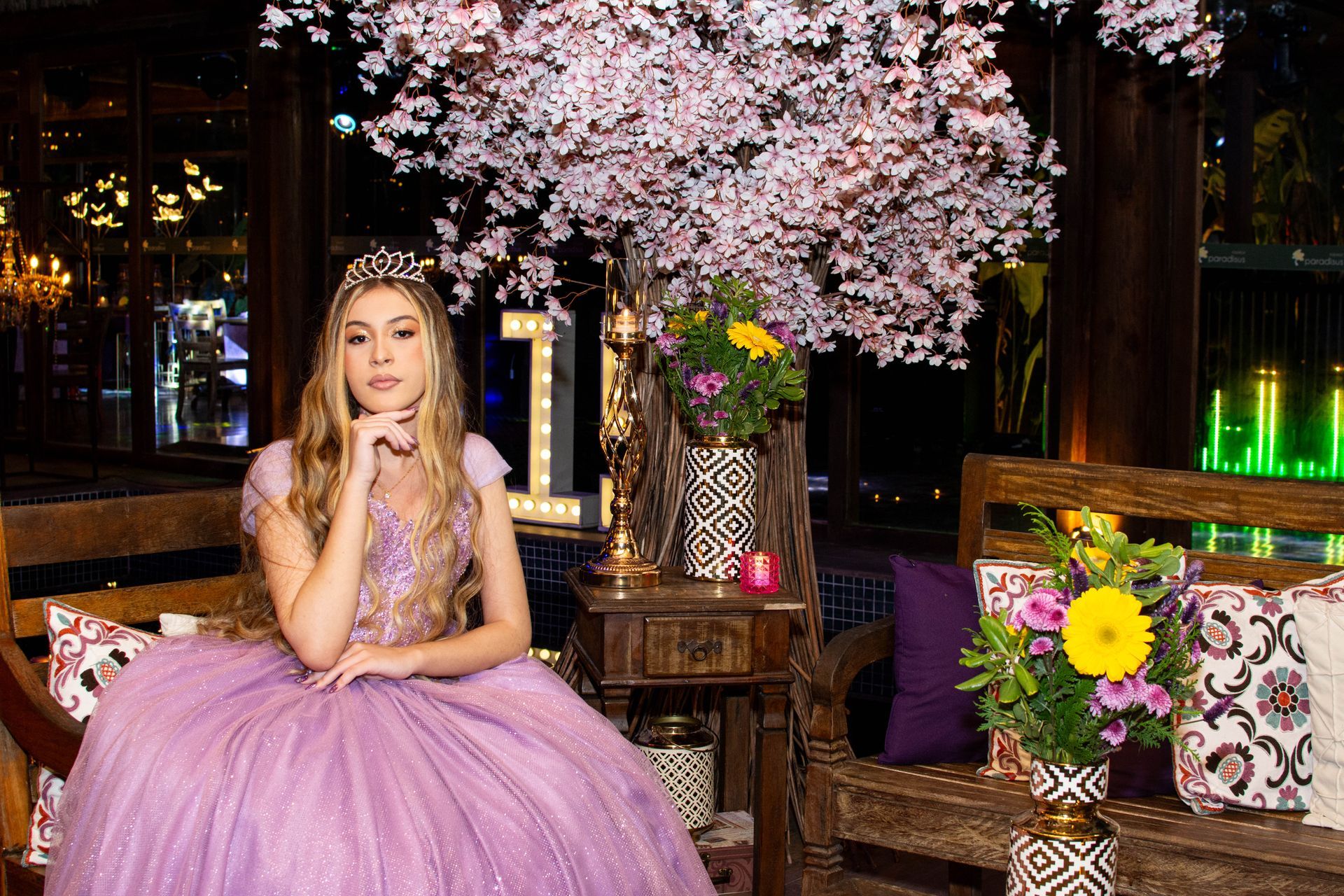 Jovem com vestido lilás e tiara, sentada perto de arranjo floral e banco decorativo.