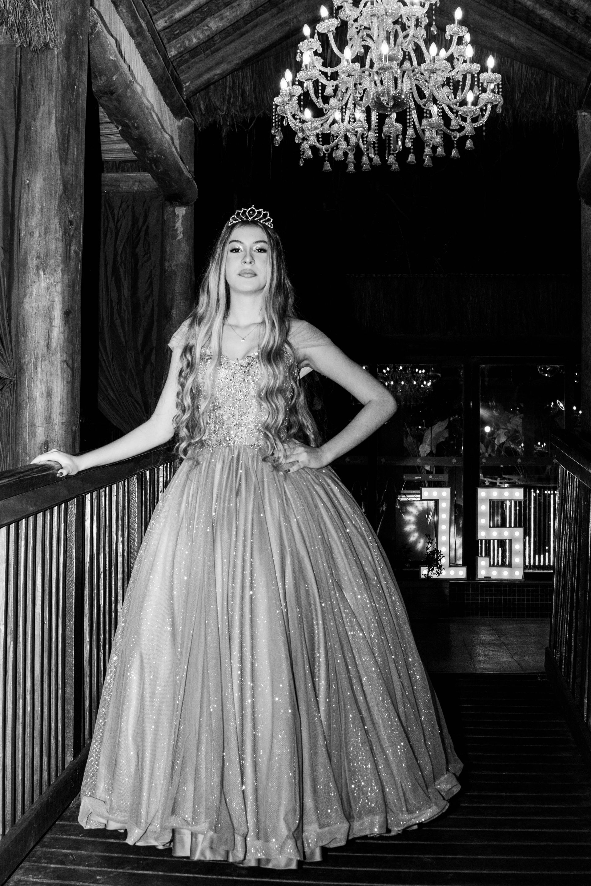 Mulher com vestido brilhante e tiara posa em uma varanda de madeira sob um lustre.