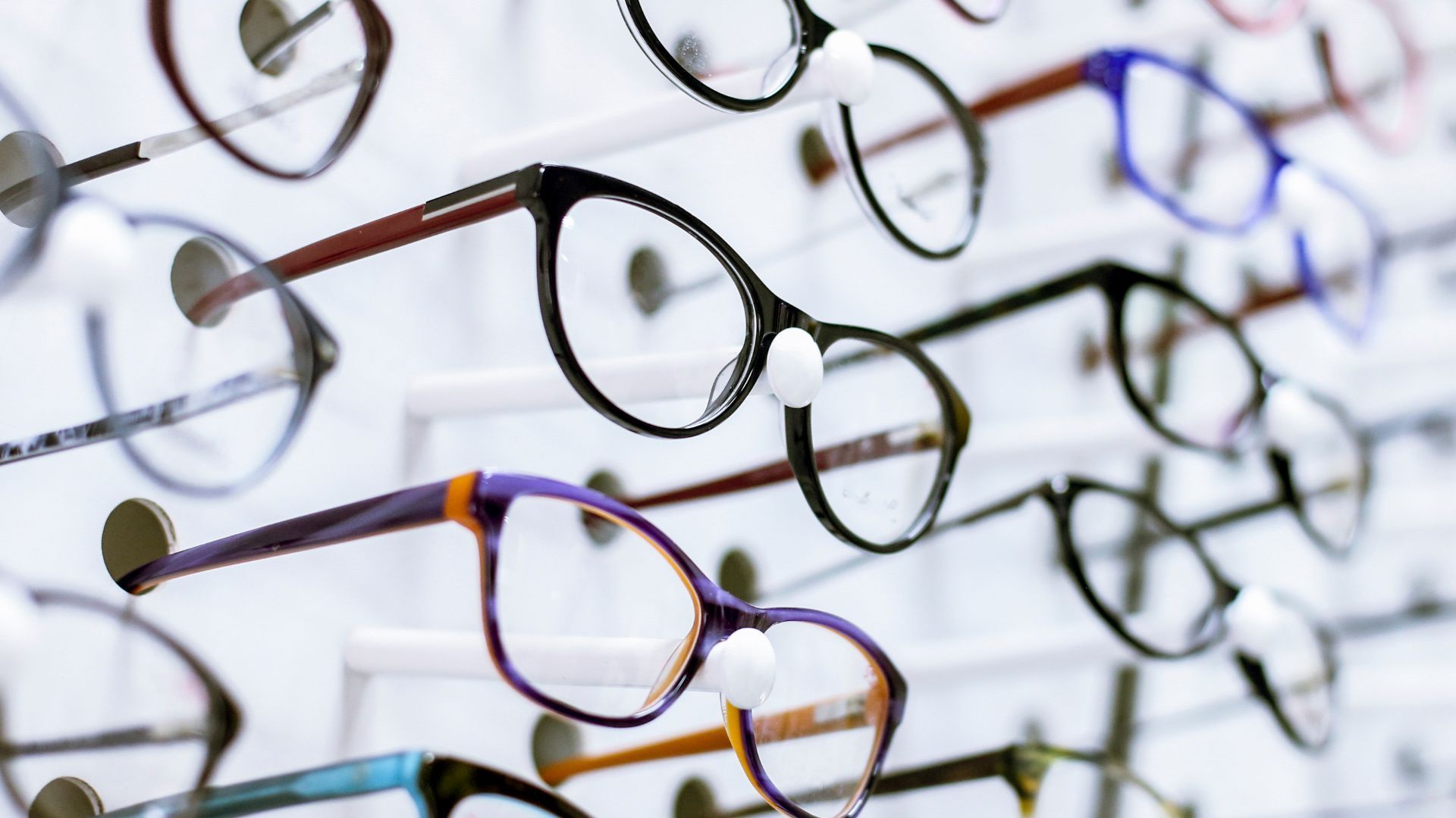 Rows of eyeglasses on display in an optician's shop. Various frame styles and colors are visible against a white wall.