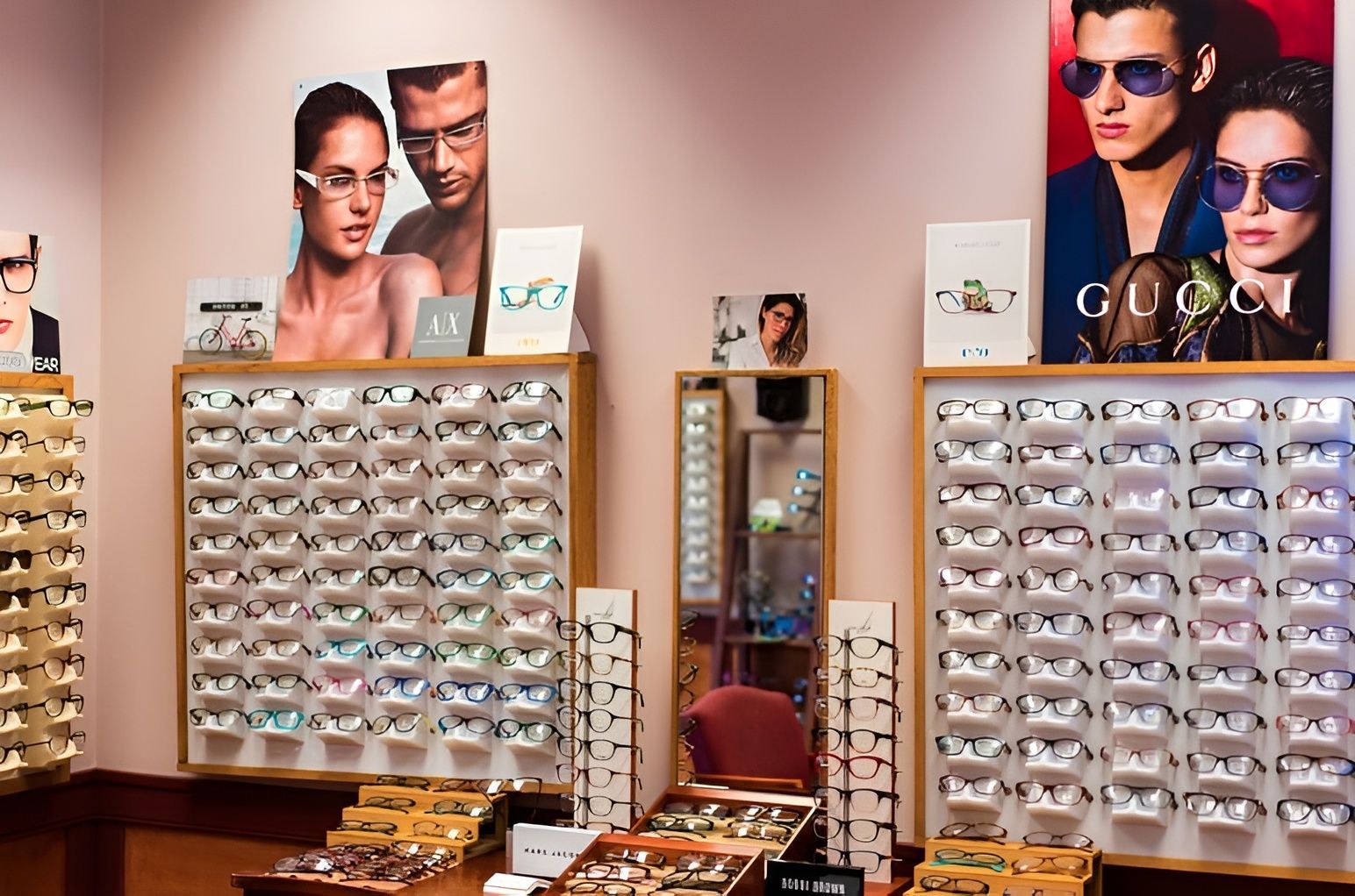 Woman with curly dark hair and glasses, smiling and holding up the glasses, wearing a light blue off-the-shoulder sweater.