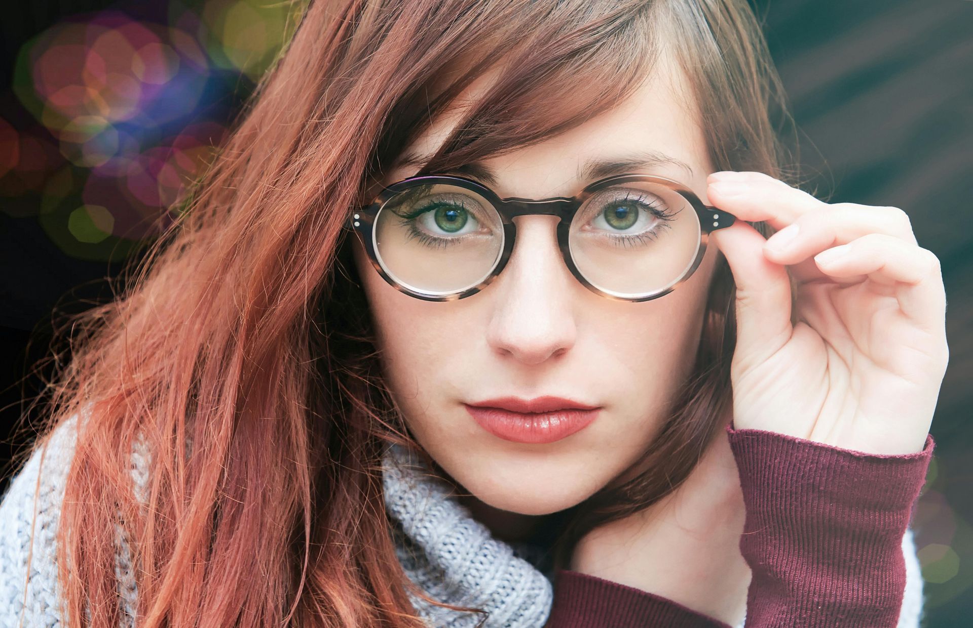 Woman with auburn hair wearing round tortoise-shell glasses, adjusting them with her right hand; looking at the viewer.