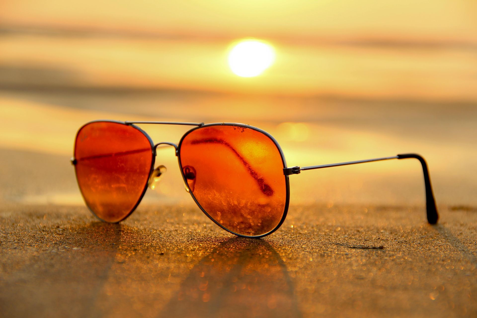 Sunglasses on sandy beach reflecting orange sunset.