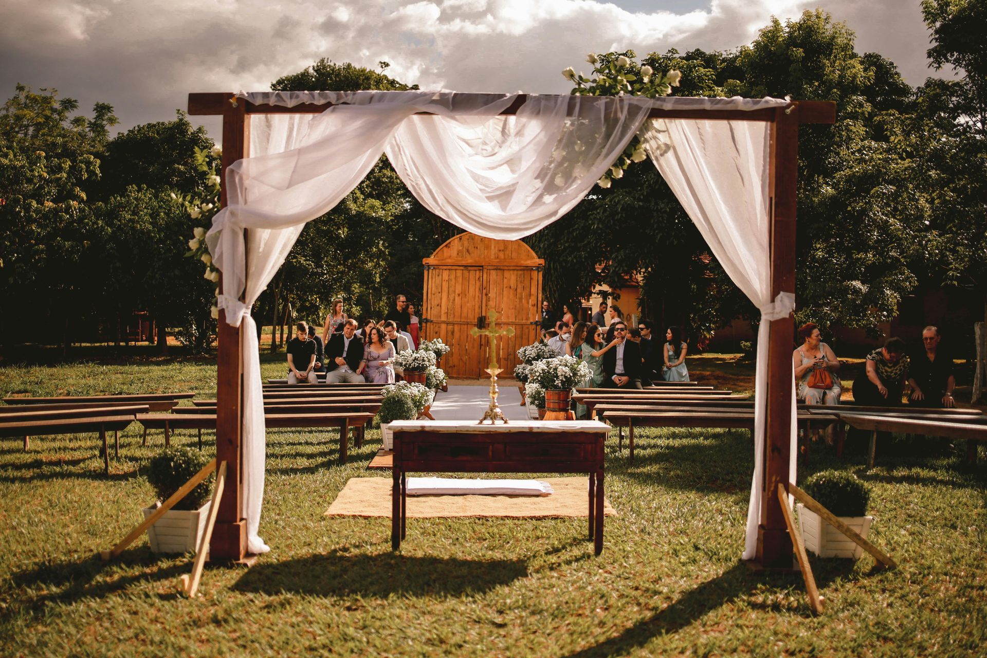 Wedding ceremony setup outdoors: wooden arch with white fabric, table, benches, and guests.