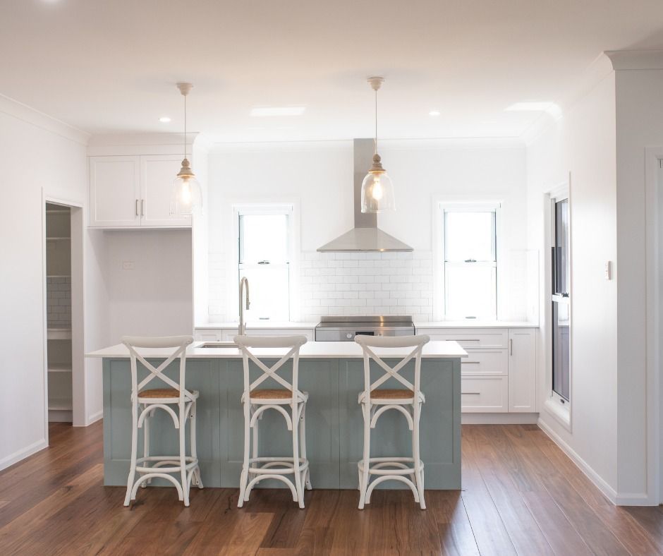 A bright, modern kitchen with a blue island, three white bar stools, wooden floors, and white cabinets and walls.