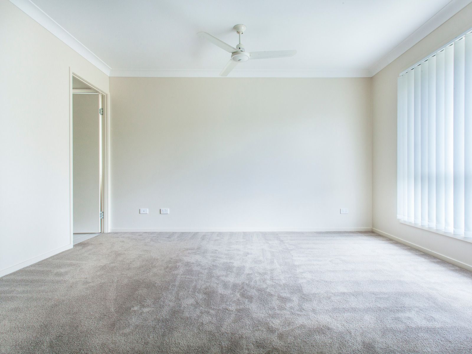 Empty room with beige walls, grey carpet, and a ceiling fan. A window with blinds is on the right.
