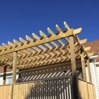 A wooden pergola with a slatted roof is attached to the side of a house under a clear blue sky.