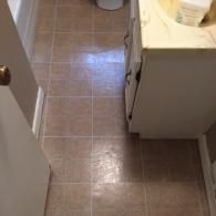 A bathroom floor with tan, square-grid vinyl tiles, a white vanity cabinet, and a door frame on the left.