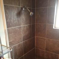 A shower stall with dark brown, square wall tiles and a silver shower head mounted on the wall.