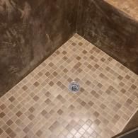 A walk-in shower floor tiled with small, square tan and brown mosaic tiles, featuring a central metal drain.