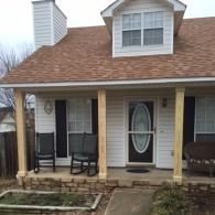 A front view of a house porch with a white door, light brown wood columns, rocking chairs, and an upper dormer window.