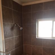A shower stall with dark brown, square wall tiles and a window on the right wall, featuring a chrome shower head.