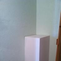 A white, box-shaped pedestal stands in the corner of a room with light green walls and a wooden door frame.