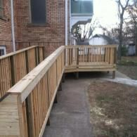 A newly constructed wooden wheelchair ramp with railings extending from the side of a brick building to a platform area.