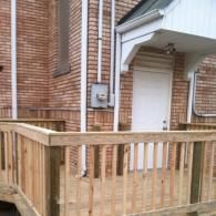 A light wood wheelchair ramp with vertical railings leads to a white door under a small roof against a brick building.