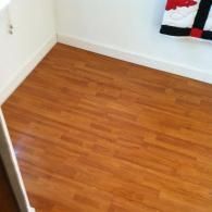 Warm-toned, wood-look laminate flooring fills the room, bordered by white baseboards with a patterned quilt in the corner.